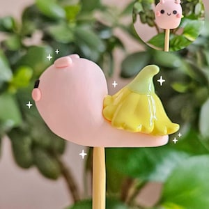 May include: A whimsical, pink snail-shaped confection with a yellow flower-like detail, mounted on a wooden stick. A smaller, similar treat is visible in a circular inset. The background features green foliage.