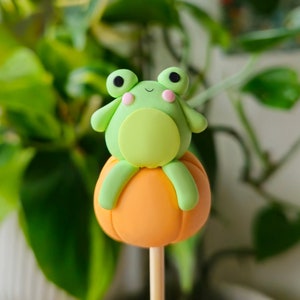 Peut inclure: Un petit gâteau en forme de citrouille orange sur un bâton en bois, surmonté d'une grenouille verte aux joues roses.