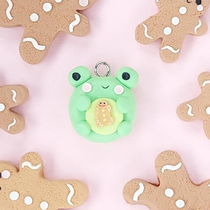 May include: A green frog figurine holding a gingerbread man cookie. The frog has a light green body with a darker green belly and white eyes. The gingerbread man is brown with white icing details. The figurine is on a pink background.