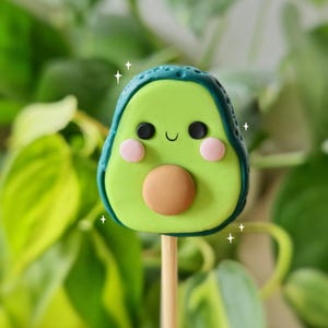 以下が含まれることがあります： 笑顔の顔、茶色の種、ピンクの頬をした緑色のアボカドの形をした置物。置物は木の棒の上にある。