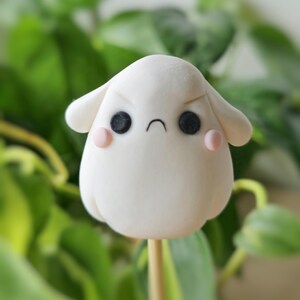 Puede incluir: Una figurita redonda de arcilla blanca con una expresión de enfado. La figurita tiene ojos negros, un ceño fruncido negro y mejillas rosadas. Está en un palo de madera.