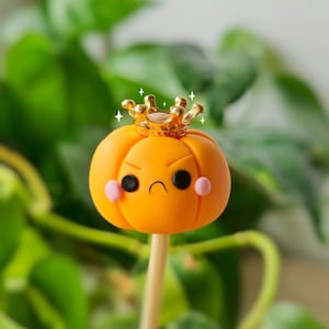 Puede incluir: Una pequeña calabaza naranja con una cara gruñona y una corona dorada en la parte superior. La calabaza está en un palo de madera.