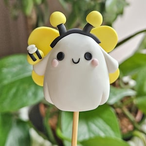 May include: A whimsical, white bee-shaped cake pop on a wooden stick. The bee has yellow wings, antennae, and a small bee on its arm. The bee has a smiling face with pink cheeks and black eyes. The background is blurred green foliage.