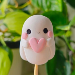 Puede incluir: Una figurita de fantasma blanca sosteniendo un corazón rosa, en un palo de madera. El fantasma tiene ojos negros y un rubor rosa en las mejillas.