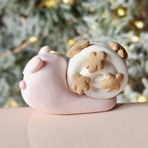 May include: A whimsical, handmade pink snail figurine with a white shell adorned with brown, gingerbread-man-shaped accents. The snail is lying down, with a small black eye and a light pink body. The background is blurred with green and yellow lights.