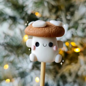 May include: A white mushroom with a brown cap and a cute cartoon face. The mushroom is on a wooden stick and is surrounded by a blurry background of a Christmas tree.