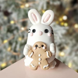 May include: A white bunny figurine with pink inner ears, black eyes, and blush-colored cheeks, holding a gingerbread man. The gingerbread man has white icing details. The bunny and gingerbread man are on a pink surface.