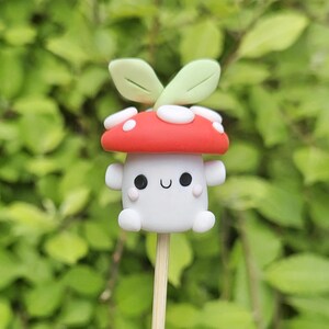 May include: A whimsical mushroom-shaped figurine with a red cap dotted with white spots, a white body, and small arms. The figurine has a smiling face and is topped with two green leaves. It is attached to a wooden stick, perfect for cake decorating.