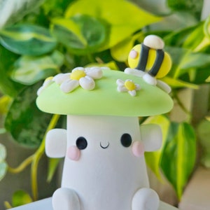 May include: A green and white mushroom figurine with a smiling face, two small pink cheeks, and a bee on top. The mushroom has white daisies on the top.