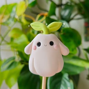May include: A white clay figurine of a cute, cartoon-like character with green leaves growing from its head. The character has a smiling face with pink cheeks and black eyes. The figurine is on a stick.