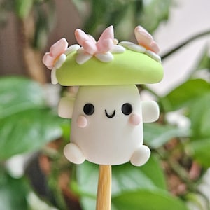 May include: A charming mushroom figurine with a smiling face, small arms, and rounded feet. The mushroom cap is light green, decorated with pink butterflies and white details. The figurine is mounted on a wooden stick, ideal for crafts or decoration.