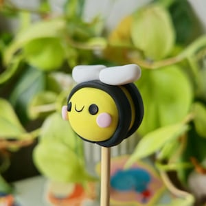 Könnte beinhalten: Eine gelbe und schwarze Biene mit einem lächelnden Gesicht und weißen Flügeln auf einem Holzstab. Die Biene ist aus Polymer Clay.