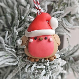 以下が含まれることがあります： 赤いサンタ帽をかぶった、白いトリムと白いポンポンが付いた、ロビンバードの形をしたお祝いのクリスマスオーナメント。鳥はピンクの胸、茶色の翼、小さな茶色の足を持っています。赤と白の紐から吊り下げられています。
