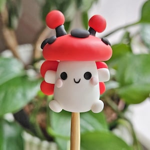 May include: A whimsical, handmade mushroom-shaped figurine on a wooden stick. The mushroom has a red cap with black accents and a white body with a smiling face and pink cheeks. The figurine is a charming decorative item.