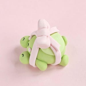 Puede incluir: Una figura de tortuga verde claro con un lazo rosa claro atado alrededor de su caparazón. La tortuga tiene pequeños ojos negros y está sobre un fondo rosa suave. El lazo añade un toque decorativo a la tortuga.