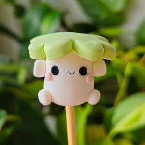 May include: A white clay figurine with a green hat and a smiling face. The figurine is on a wooden stick.