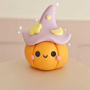 May include: A small orange pumpkin figurine with a smooth matte finish, featuring a smiling face with black circular eyes and pink cheeks. It wears a light purple pointed hat decorated with a yellow star, a yellow crescent moon, and yellow dots.