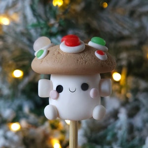 May include: A white and brown clay mushroom ornament with a smiling face and red, green, and white candy decorations on top. The ornament is on a wooden stick.