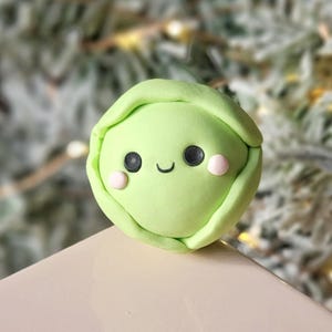 May include: A light green, round, handcrafted sculpture of a smiling brussels sprout. The sprout has black eyes, a curved mouth, and pink cheeks. The background is a blurred image of a Christmas tree.