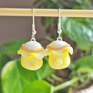May include: A pair of yellow and white mushroom-shaped earrings with silver hooks. The earrings have a detailed design with a small, circular accent on the front. The background is blurred, with green foliage.