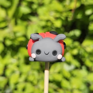 May include: A whimsical ladybug-shaped cake pop on a wooden stick. The ladybug has a red body with black spots, gray head, and small white cheeks. The background is a blurred green foliage.
