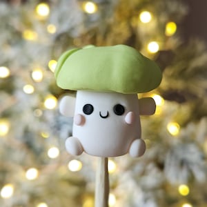 May include: A whimsical, handmade mushroom figurine with a light green cap and a smiling face. The mushroom has small arms and legs, and is mounted on a wooden stick. The background is blurred with bokeh lights.