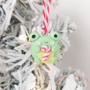 May include: A green frog Christmas ornament holding a candy cane. The ornament is made of polymer clay and has a red and white string for hanging.