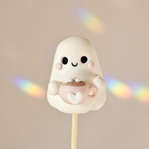 May include: A white ghost-shaped cake pop with black eyes, a curved mouth, and pink cheeks. The ghost holds a pink teacup with a heart design. The cake pop is on a wooden stick, set against a neutral background.