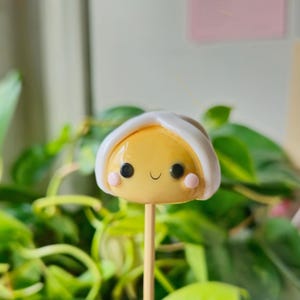 Puede incluir: Una figurita de arcilla amarilla y blanca de una cara sonriente con un sombrero blanco. La figurita está en un palo de madera.