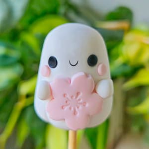 May include: A white, cartoon-style figure with black eyes, pink cheeks, and a smiling mouth, holding a pink flower. The character is on a wooden stick, possibly a cake pop. The background features blurred green leaves.