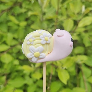 Puede incluir: Una caprichosa figura de caracol hecha a mano sobre un palito de madera. La concha del caracol es amarilla con detalles de margaritas blancas, y el cuerpo es rosa. El caracol tiene ojos negros, sobre un fondo verde borroso.