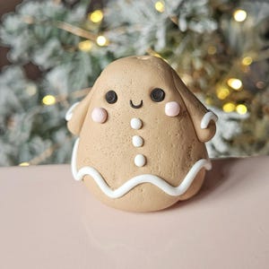 May include: A gingerbread man ornament with a smiling face, black eyes, and pink cheeks. The brown ornament has white icing details and three white buttons down the front. The background is a blurred Christmas tree.