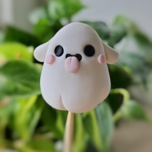 May include: A white ghost-shaped figurine with black eyes, a pink tongue sticking out, and pink cheeks. The figurine is on a wooden stick.