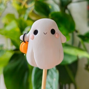 May include: A white ghost figurine with black eyes and a small pink blush on its cheeks. The ghost is holding a small orange pumpkin in its hand and is attached to a wooden stick.