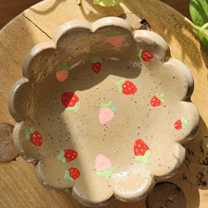 May include: A handmade ceramic dish with a scalloped edge, decorated with painted strawberries. The strawberries are red and pink with green leaves, scattered across the beige surface. The dish is on a wooden surface.