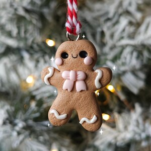 May include: A gingerbread man Christmas ornament made of brown clay with white icing details and a pink bow. The ornament is hanging from a red and white striped string.
