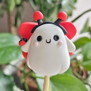 May include: A whimsical, white figurine resembling a ladybug, with red wings and antennae, and a smiling face. The figurine is mounted on a wooden stick, perfect for cake decorating or as a unique craft supply.