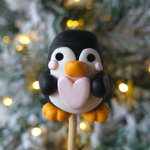 Puede incluir: Una figurita de pingüino blanco y negro sosteniendo un corazón rosa, con patas naranjas y un palo de madera, sobre un fondo borroso de un árbol de Navidad con luces blancas.