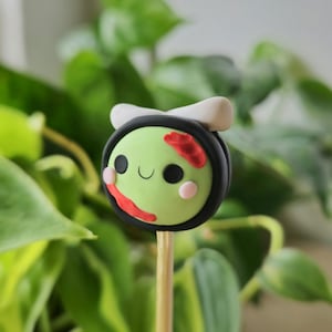 Puede incluir: Un cake pop zombie verde y negro con una diadema blanca y detalles rojos y negros en la cara. El cake pop está en un palo de madera.