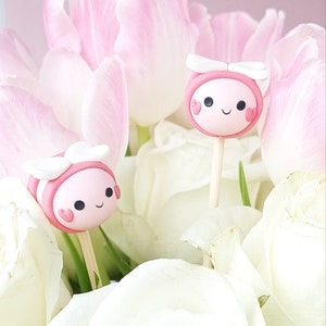 May include: Two pink and white bee-shaped cake pops with heart-shaped eyes and wings. The cake pops are on sticks and are surrounded by white flowers.