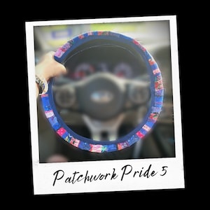 以下が含まれることがあります： カラフルなパッチワークのデザインが施された青いステアリングホイールカバー。カバーはピンク、ブルー、ホワイトのストライプになっています。画像には「Patchwork Pride 5」というテキストが表示されています。