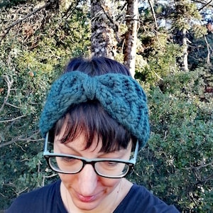 May include: A teal blue knitted headband with a bow detail. The headband is worn by a person with short, dark hair and glasses.