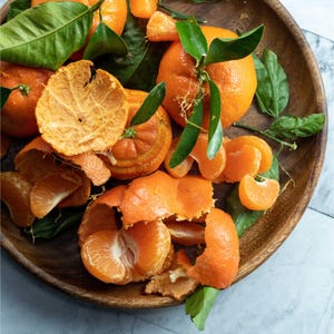 May include: A wooden bowl filled with fresh, ripe tangerines. Some are whole with green leaves, others are peeled, revealing juicy segments. The vibrant orange fruit and peel contrast with the dark wood and green leaves.