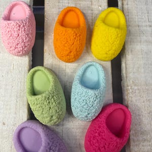 Könnte beinhalten: Sieben farbenfrohe, strukturierte Seifen in Pantoffelform in den Farbtönen Rosa, Orange, Gelb, Grün, Blau, Lila und Fuchsia. Die Seifen sind auf einer hellen Holzoberfläche angeordnet.