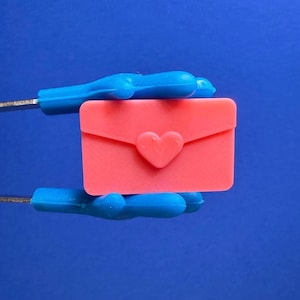 May include: A small, coral-colored envelope with a heart-shaped clasp. The envelope is held against a vibrant blue background. The heart clasp is centered on the front of the envelope.