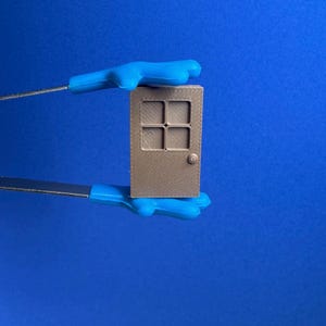 May include: A small, tan-colored miniature door with a four-pane window design and a round doorknob. The door is held by blue-gloved hands against a solid blue background. The door appears to be a 3D-printed item.