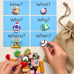 May include: Six light blue rectangular cards labeled How?, What?, Who?, Why?, Where?, and When? with attached miniature plastic charms including a mixer, snail, rocket, accordion, sports shop, and baseball. A pile of assorted colorful charms sits nearby.