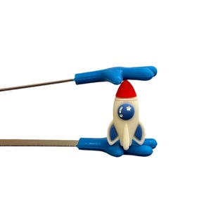 May include: A rocket-shaped bottle stopper with a red top, white body, and blue accents. The stopper is held by blue-handled tongs with silver metal prongs. The rocket has a star and moon design on the side.