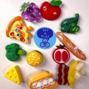 May include: A collection of miniature food-shaped charms including purple grapes, a red apple, green broccoli, a pizza slice, a yellow burger, a baguette, a cheese wedge, bacon, a banana, and a carton of eggs with faces, surrounding a blue Say & Play logo.