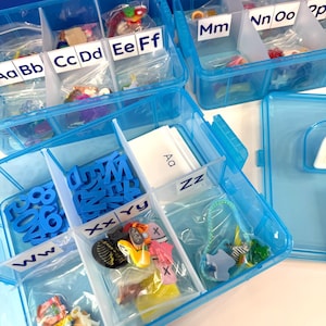 Könnte beinhalten: Drei blaue Kunststoff-Aufbewahrungsboxen mit mehreren Fächern, die jeweils mit Buchstaben des Alphabets von Aa bis Zz beschriftet sind. Die Fächer enthalten kleine Plastiktüten mit verschiedenen Miniaturobjekten und blauen Buchstabenausschnitten.